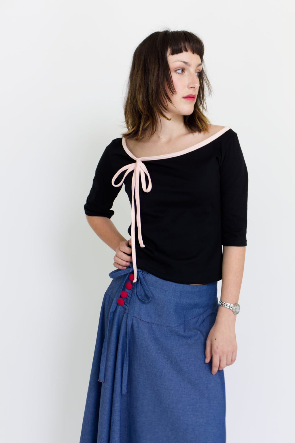 A young woman wearing a black and baby pink boat neck top with a ¾ sleeve and bow, paired with a denim blue chambray maxi skirt, in front of a white back ground
