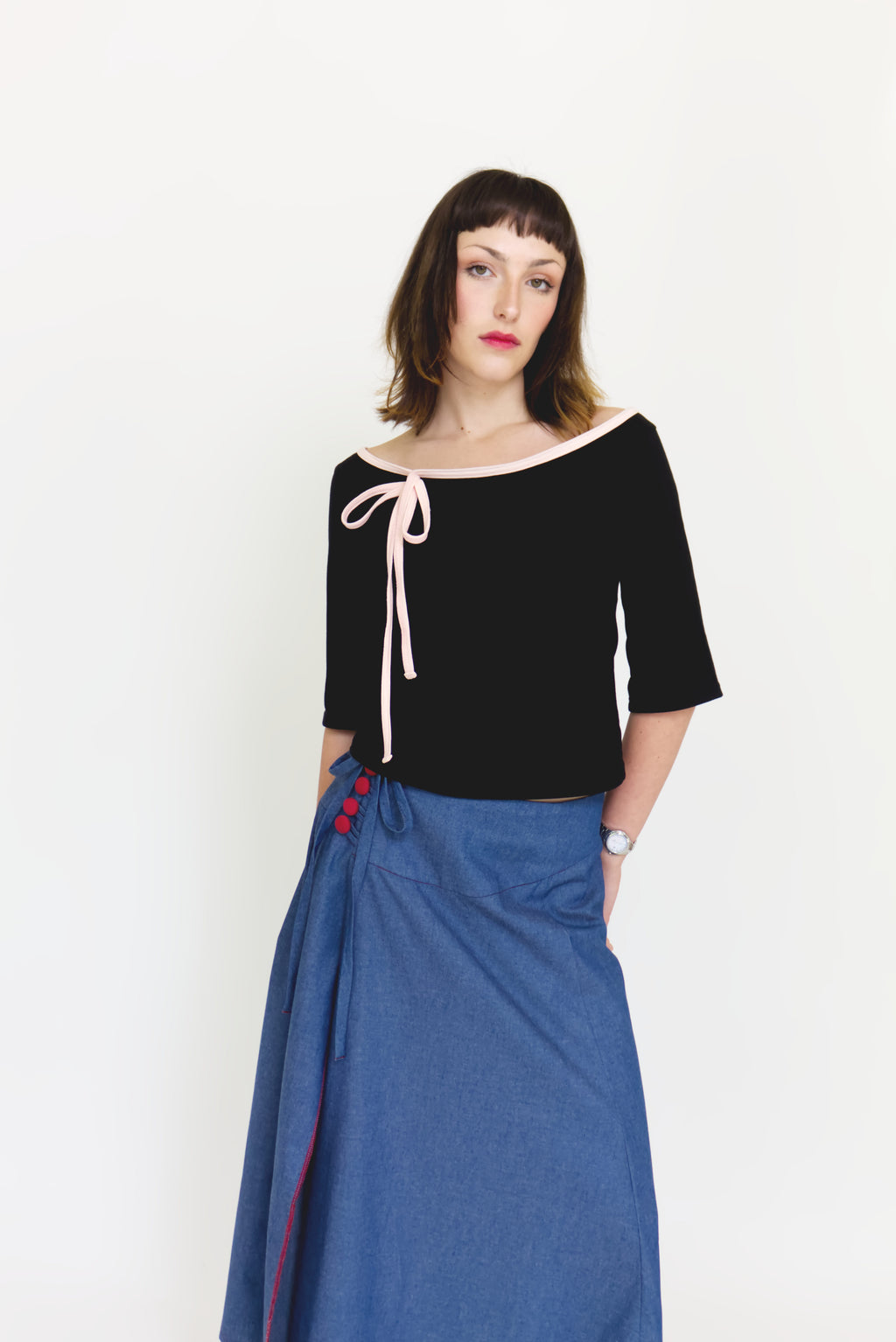 A young woman wearing a black and baby pink boat neck top with a ¾ sleeve and bow, paired with a denim blue chambray maxi skirt, in front of a white back ground