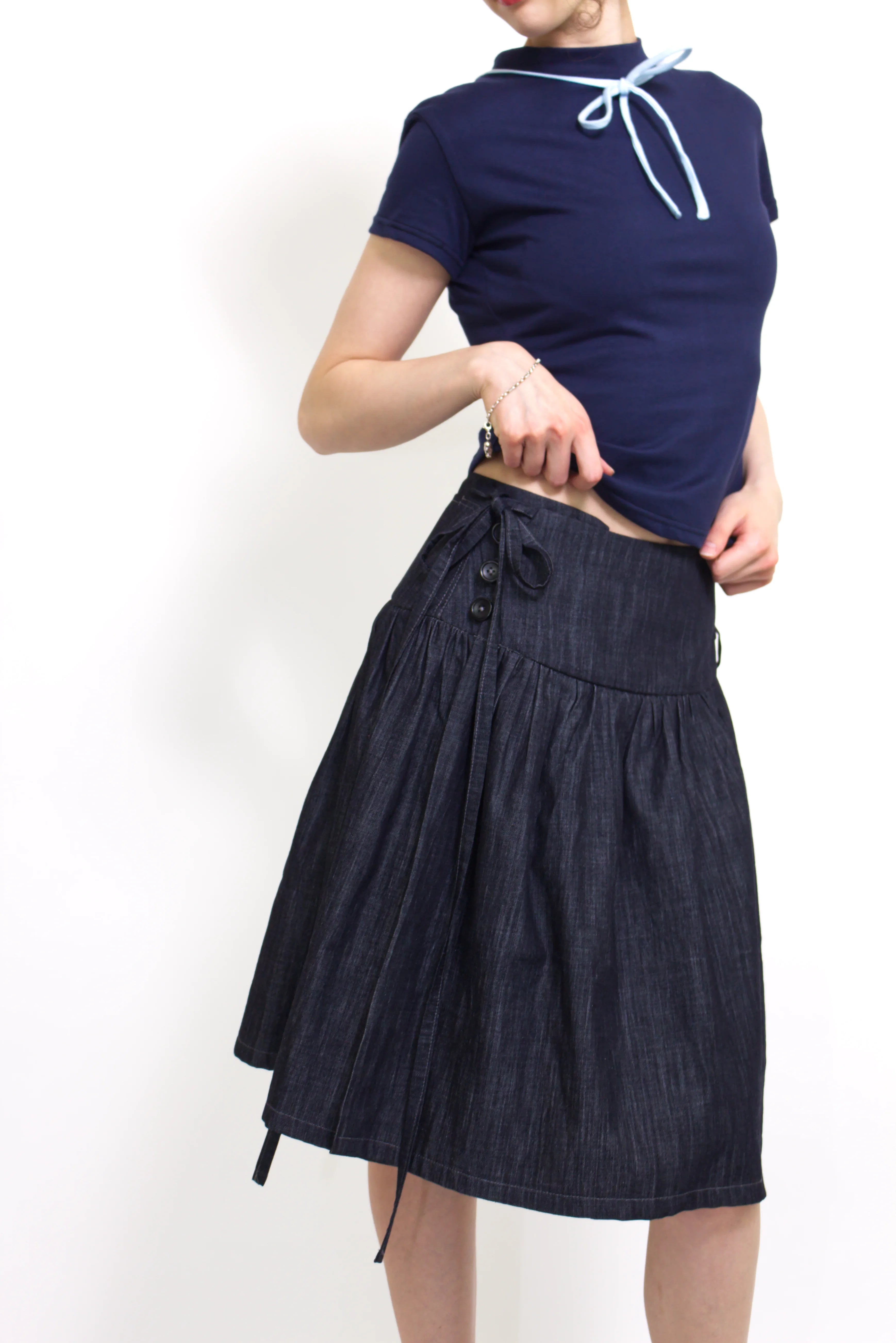 Woman wearing a navy and baby blue short sleeve top with a mock neck and blue bow with an indigo denim knee length gathered wrap skirt against a white background