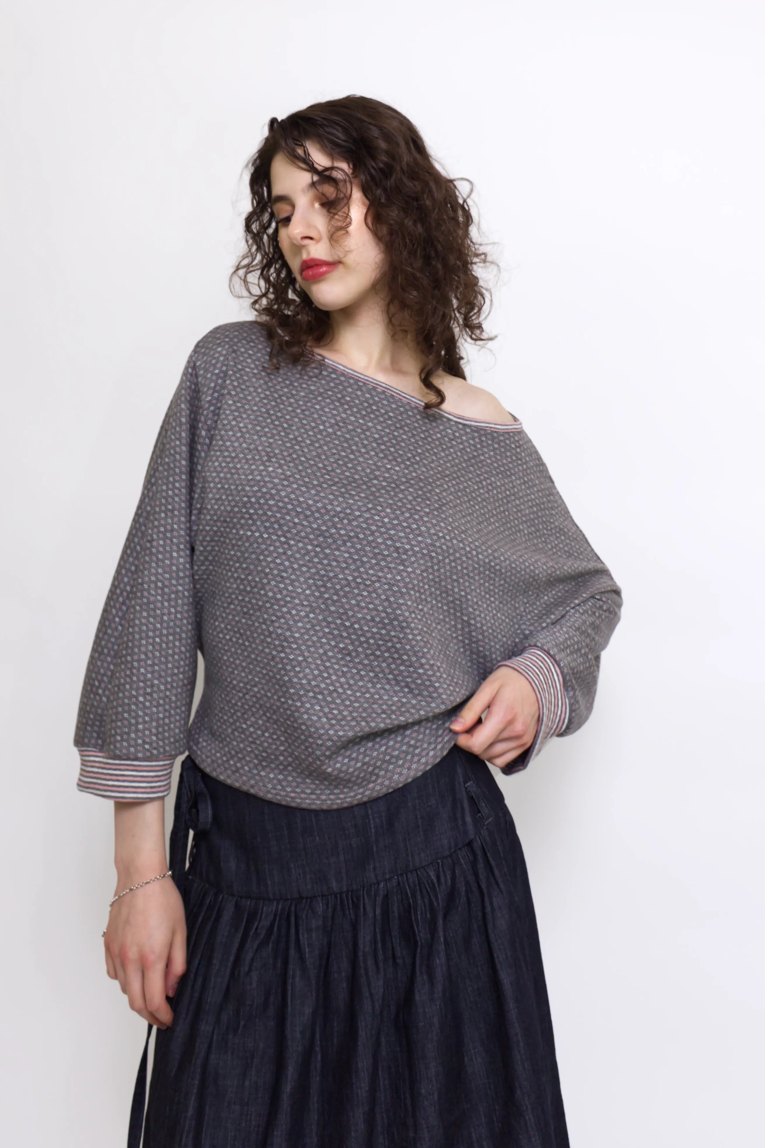 young woman wearing a grey, pink and white patterned oversized cotton boat neck pullover top, with long sleeves on a white background