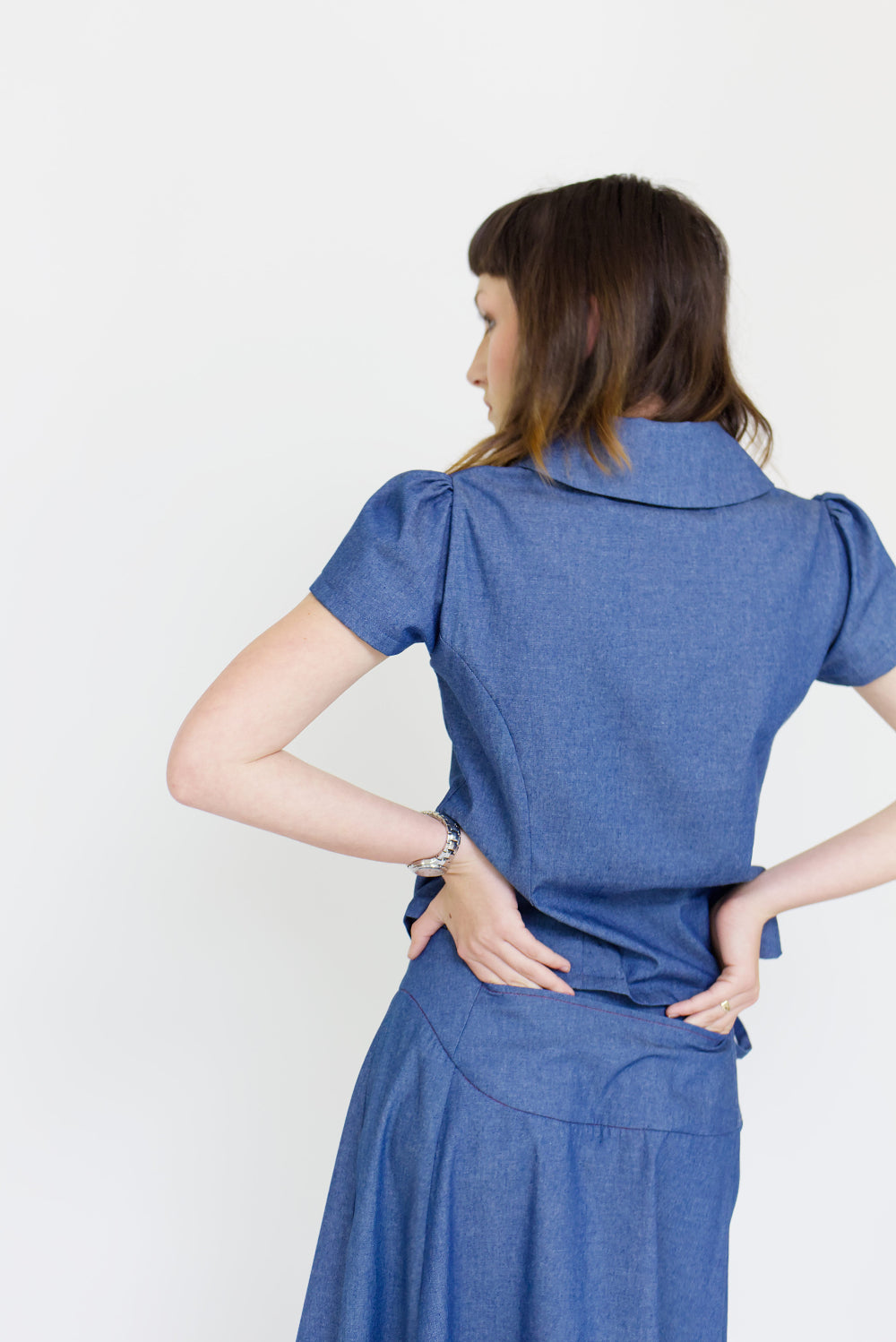 Young woman in a matching blue chambray cap sleeve, peter pan collar, princess line blouse and asymmetrical, low rise maxi warp skirt with a split, both with red buttons, in front of a white back ground
