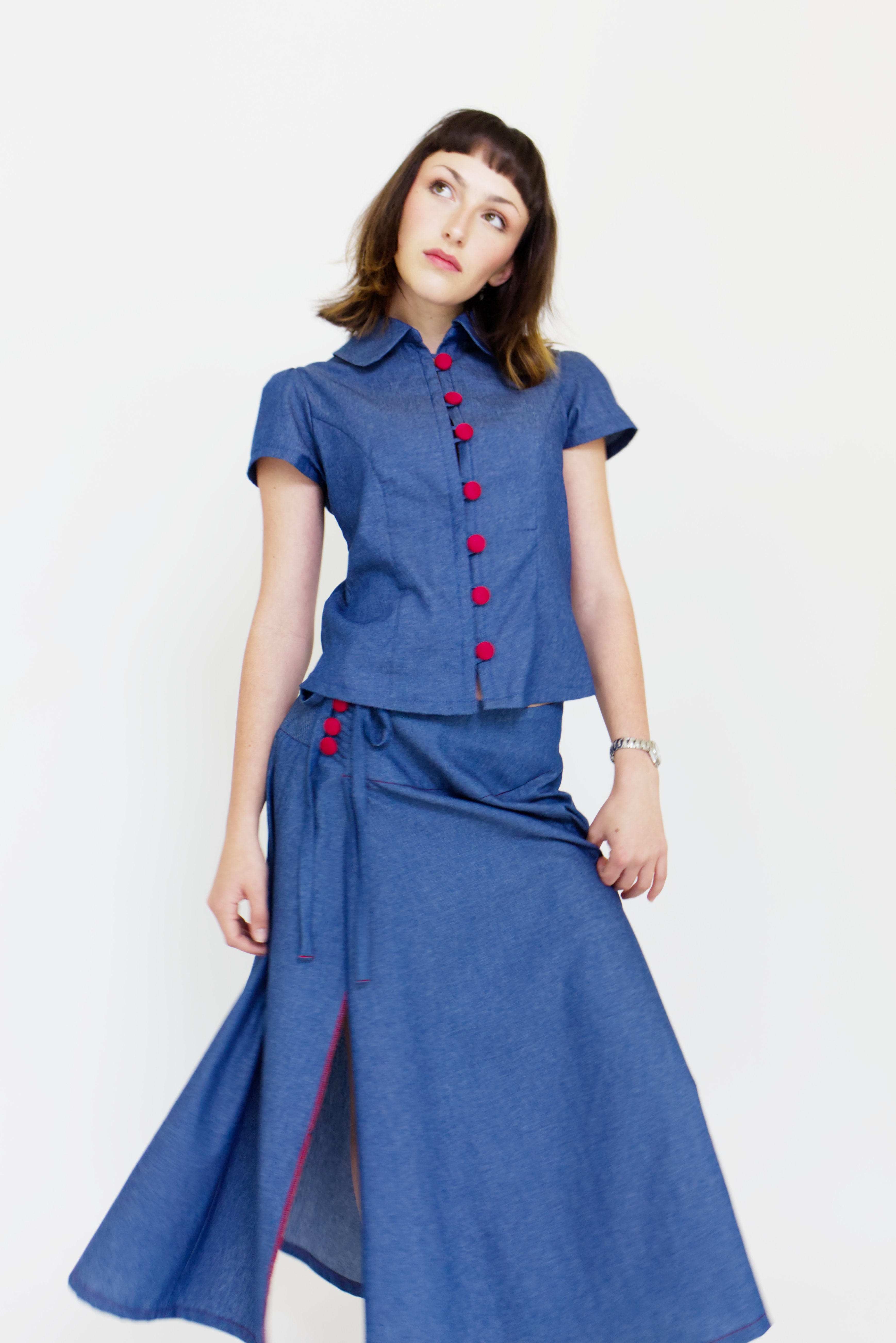 Young woman in a matching blue chambray cap sleeve, peter pan collar, princess line blouse and asymmetrical, low rise maxi warp skirt with a split, both with red buttons, in front of a white back ground
