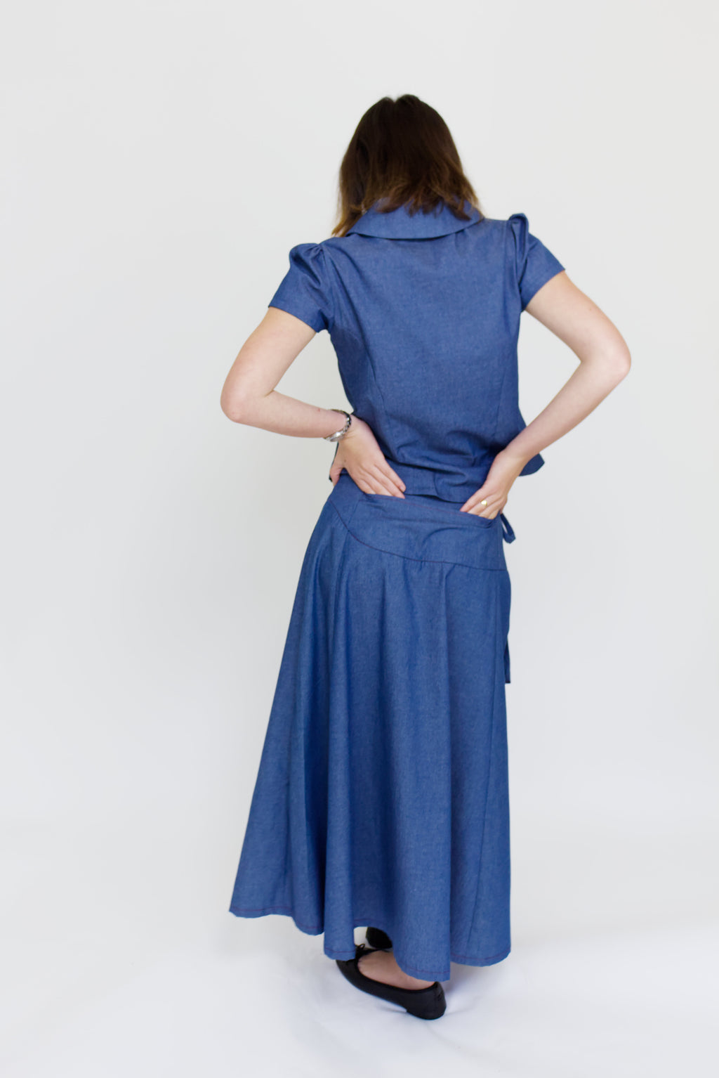 Young woman in a matching blue chambray cap sleeve, peter pan collar, princess line blouse and asymmetrical, low rise maxi warp skirt with a split, both with red buttons, in front of a white back ground
