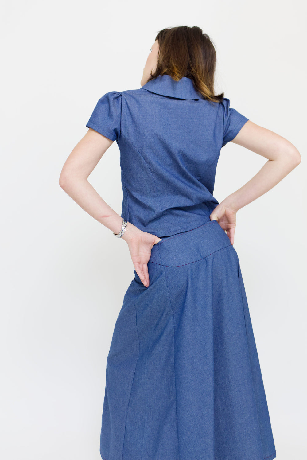 Young woman in a matching blue chambray cap sleeve, peter pan collar, princess line blouse and asymmetrical, low rise maxi warp skirt with a split, both with red buttons, in front of a white back ground
