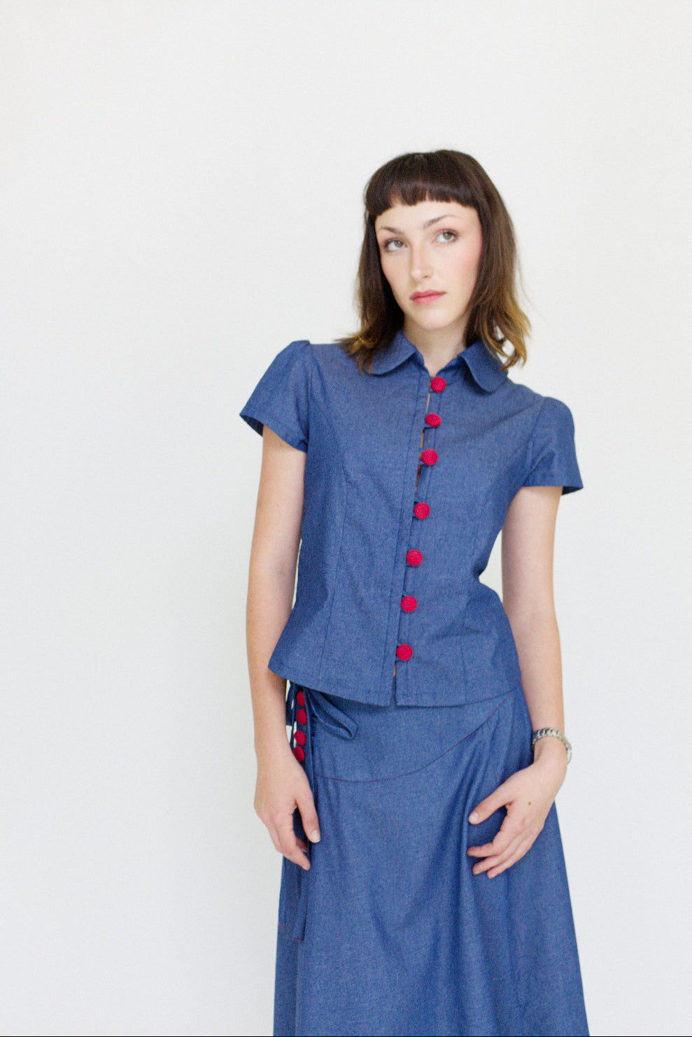 Young woman in a matching blue chambray cap sleeve, peter pan collar, princess line blouse and asymmetrical, low rise maxi warp skirt with a split, both with red buttons, in front of a white back ground
