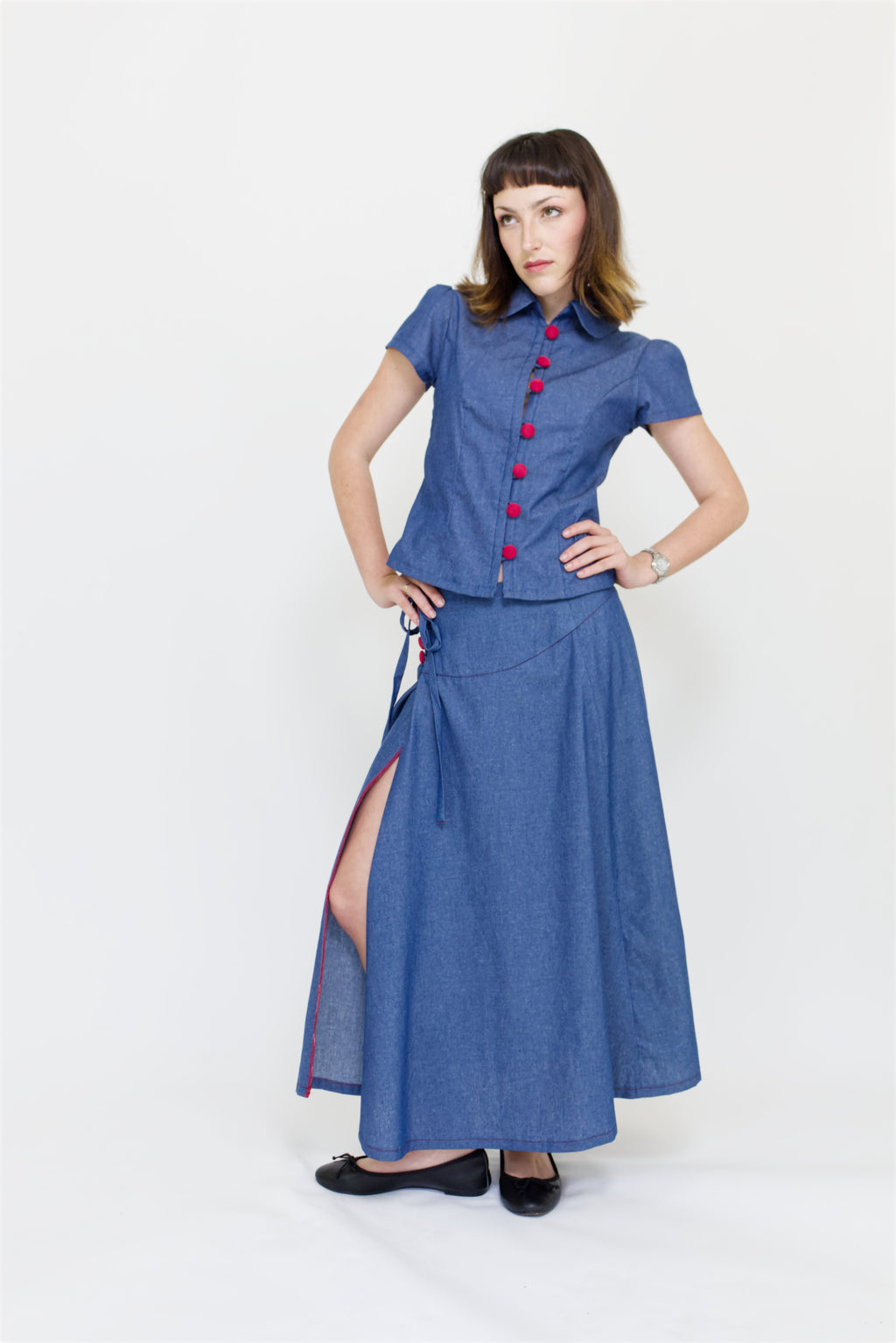 Young woman in a matching blue chambray cap sleeve, peter pan collar, princess line blouse and asymmetrical, low rise maxi warp skirt with a split, both with red buttons, in front of a white back ground
