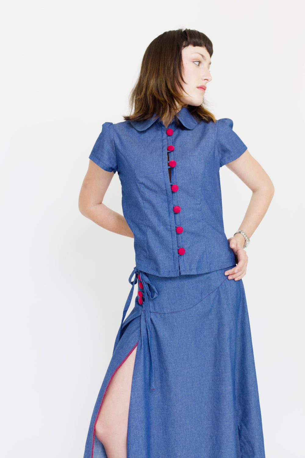 Young woman in a matching blue chambray cap sleeve, peter pan collar, princess line blouse and asymmetrical, low rise maxi warp skirt with a split, both with red buttons, in front of a white back ground
