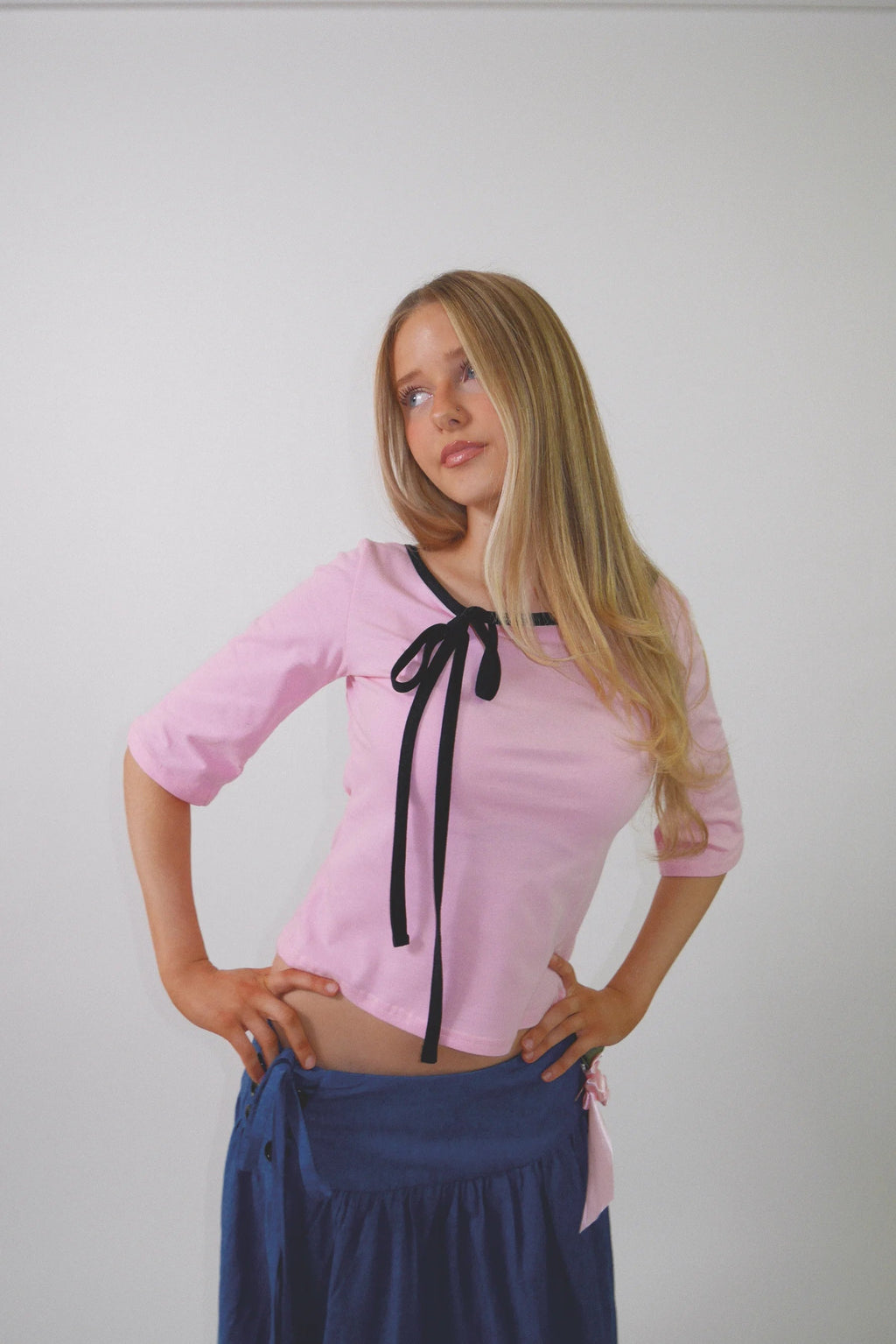 A woman with long blonde hair posing in a light pink short-sleeve top with a black ribbon tie at the collar, paired with a navy blue skirt, against a plain light gray background.