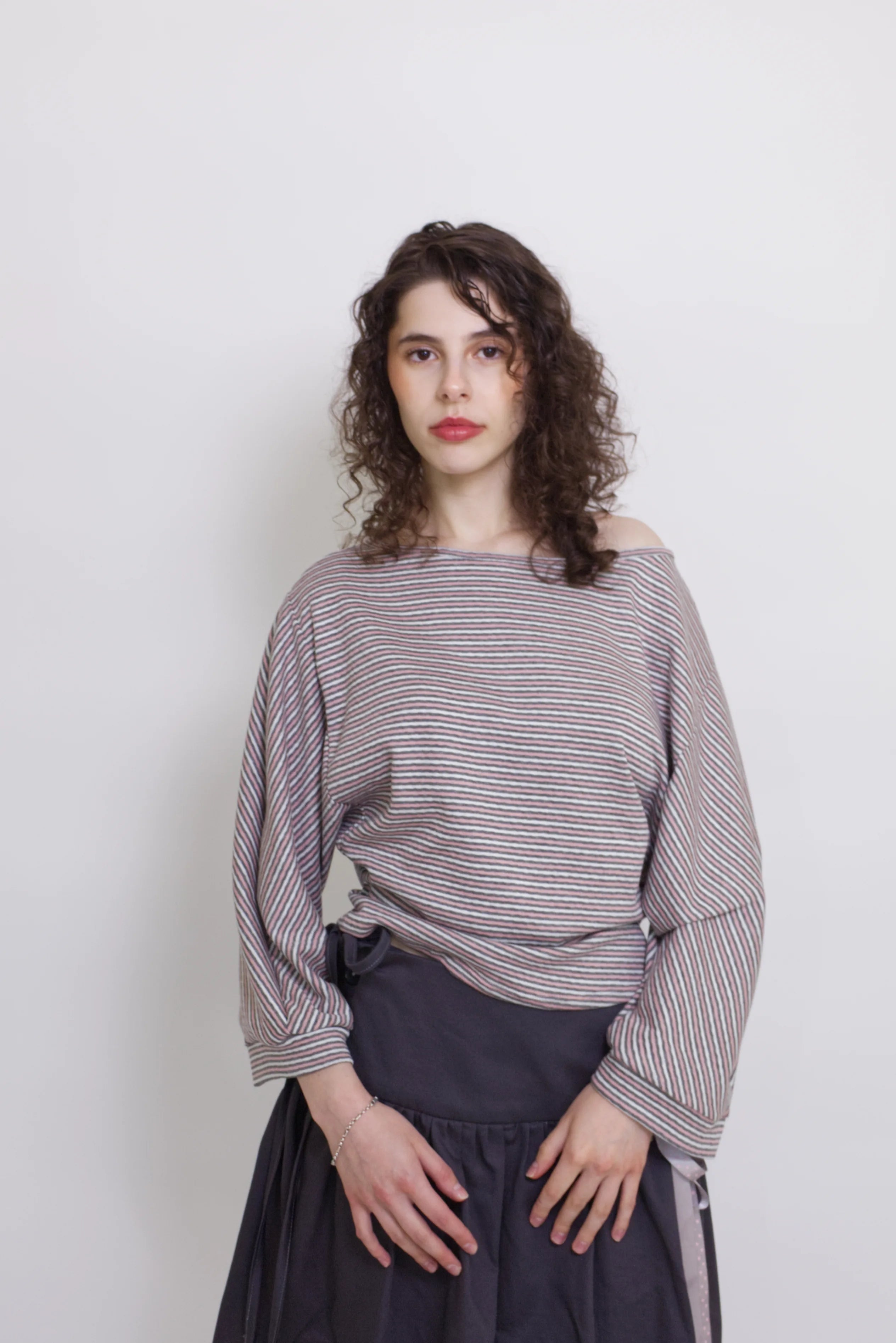 young woman wearing a pink striped oversized cotton boat neck pullover top, with long sleeves on a white background